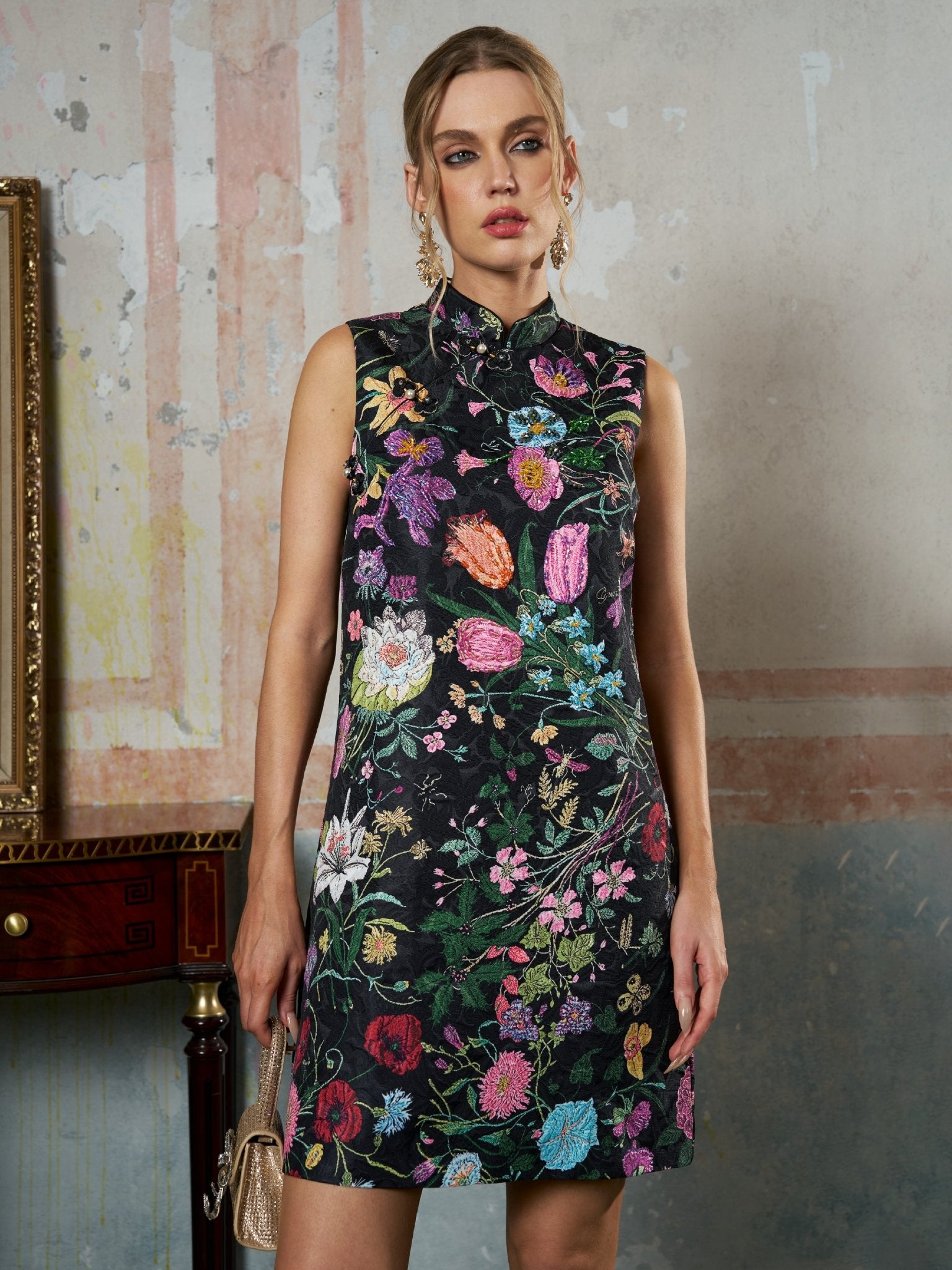 A woman stands indoors wearing the Social Queen Colorful Floral Mandarin Collar Dress, holding a small metallic purse, with a painted wall and wooden table in the background.