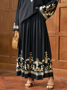 Wearing the Social Queen Contrast Embroidered A-Line Skirt, a person stands on a wooden floor by a paneled wall, holding a wicker handbag and wearing gold heeled sandals.