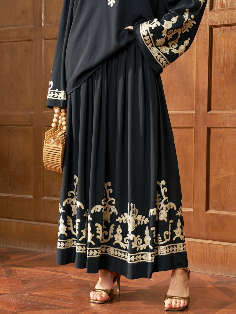 Wearing the Social Queen Contrast Embroidered A-Line Skirt, a person stands on a wooden floor by a paneled wall, holding a wicker handbag and wearing gold heeled sandals.