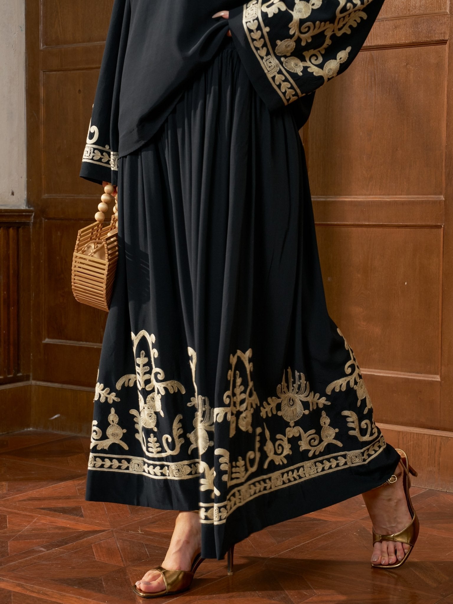 Wearing the Social Queen Contrast Embroidered A-Line Skirt, a person stands in heeled sandals with a wooden bag on a wooden floor against a wood-paneled wall.