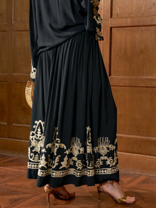A person stands on a wooden floor in the Social Queen Contrast Embroidered A-Line Skirt, paired with brown heeled sandals and holding a woven handbag, next to a wooden wall.