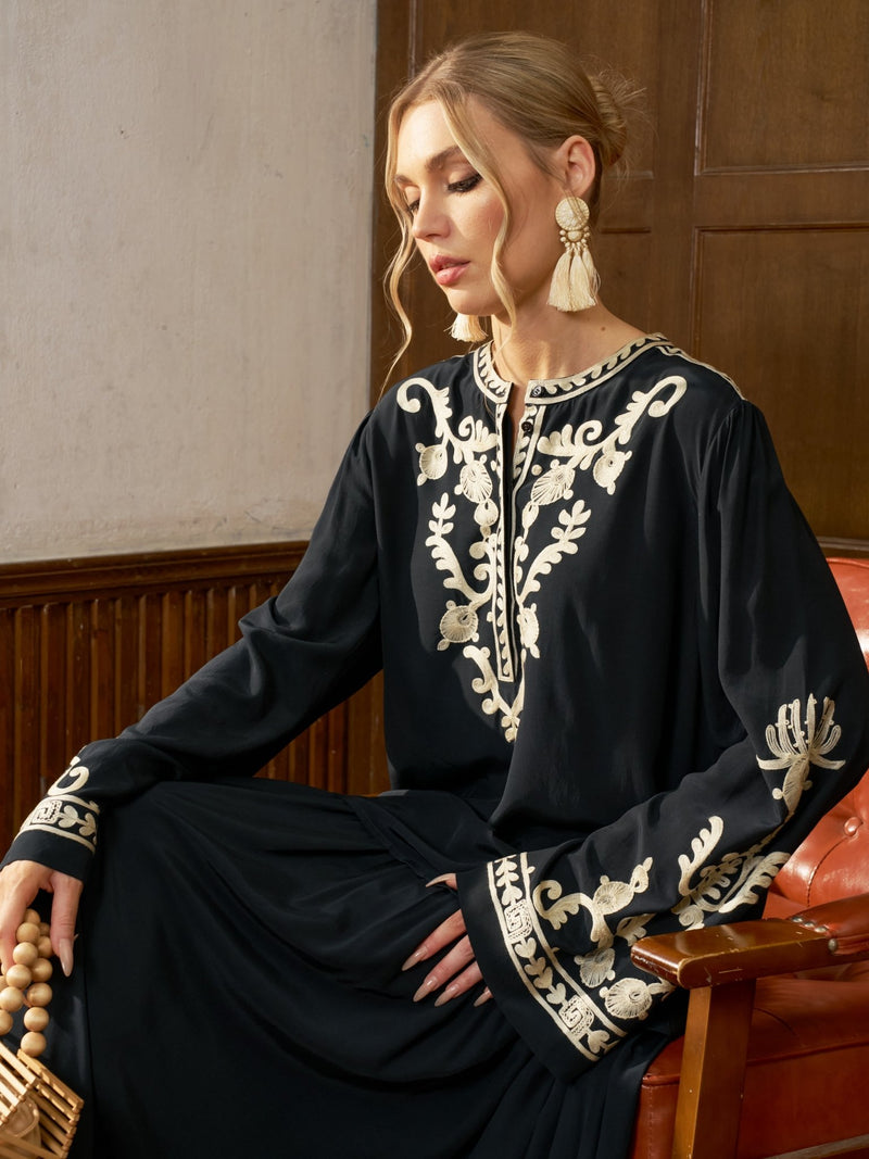 A woman seated in an armchair wears the Social Queen Contrast Embroidered Relaxed Blouse, large tassel earrings, and holds wooden beads.