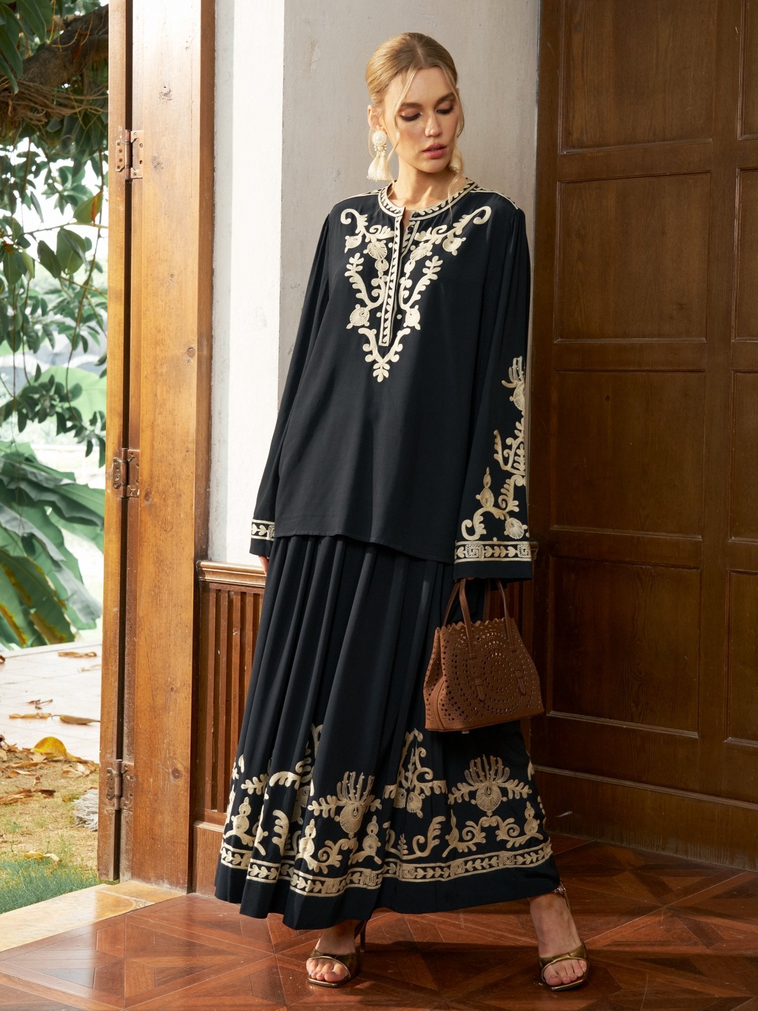 A woman stands indoors wearing the Social Queen Contrast Embroidered Relaxed Blouse, holding a brown handbag, with a wooden door and greenery in the background.