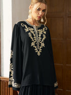 A woman stands indoors near wooden panels, wearing the Social Queen Contrast Embroidered Relaxed Blouse—a black chiffon blend long-sleeve top with intricate contrast embroidery.