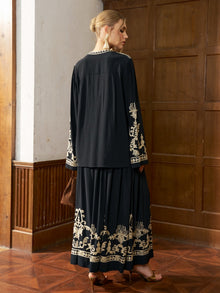 A woman stands indoors, facing away, wearing the Social Queen Contrast Embroidered Relaxed Blouse styled with a matching jacket and long black skirt. She holds a brown bag and wears heeled sandals for a chic look with a relaxed silhouette.