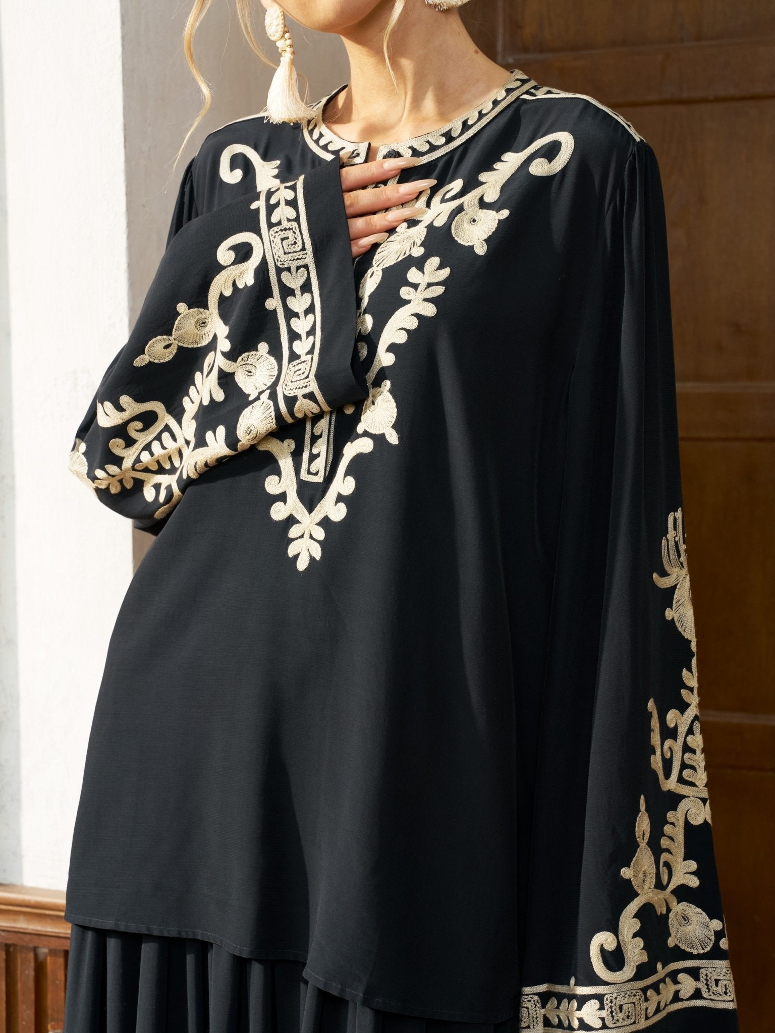 A person wears the Social Queen Contrast Embroidered Relaxed Blouse, a loose black top with contrast embroidery on the sleeves, neckline, and hem, standing against a wooden door and white wall.