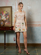 A woman wears the Social Queen Contrast Floral Embroidered Mini Dress—a fitted, light-toned piece—indoors while holding a small purse. Behind her is a wooden table with a framed floral painting.