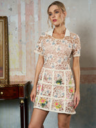 A woman wears the Social Queen Contrast Floral Embroidered Mini Dress, featuring short sleeves, a white collar, and a fitted straight silhouette. Her hair is in an updo with earrings; a wooden table and framed artwork are behind her.