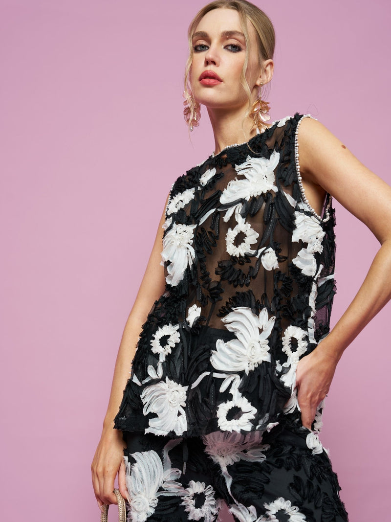 A woman poses against a pink background in the Social Queen Contrast Floral Sheer Top— a sleeveless black mesh top with bold white and black floral patterns, styled with matching pants.