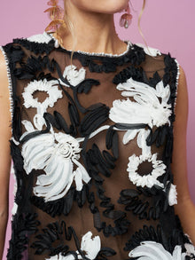 A model wears the Social Queen Contrast Floral Sheer Top, a sleeveless black mesh with bold white and black floral embroidery, set against a pink background.