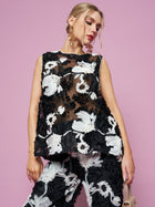A woman poses against a pink background wearing the Social Queen Contrast Floral Sheer Top, a sleeveless black mesh piece with bold white and black floral embroidery.