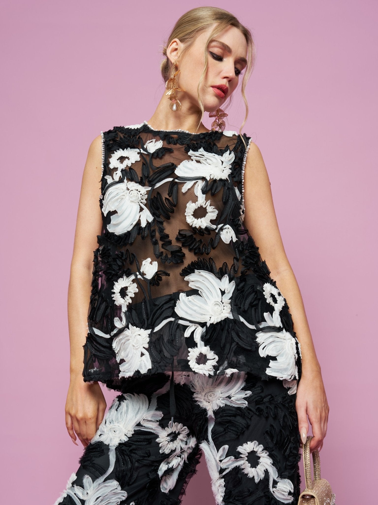 A woman poses against a pink background wearing the Social Queen Contrast Floral Sheer Top, a sleeveless black mesh piece with bold white and black floral embroidery.