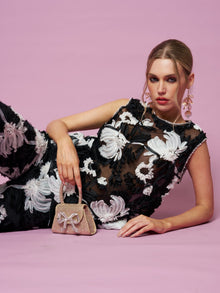 Model reclines on a pink background wearing the Social Queen Contrast Floral Sheer Top, paired with a small pink handbag adorned with a bow.