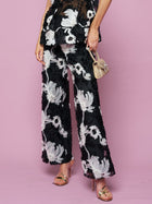 Model wears Social Queen’s Contrast Floral Sheer Wide-Leg Pants with a floral top, gold high-heeled sandals, and a metallic handbag against a pink background.