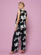 A woman poses against a pink backdrop in Social Queen's Contrast Floral Sheer Wide-Leg Pants, styled with a sleeveless black top, nude heels, and gold earrings.