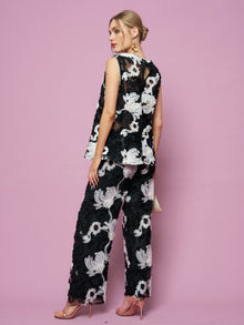 A woman poses against a pink backdrop in Social Queen's Contrast Floral Sheer Wide-Leg Pants, styled with a sleeveless black top, nude heels, and gold earrings.