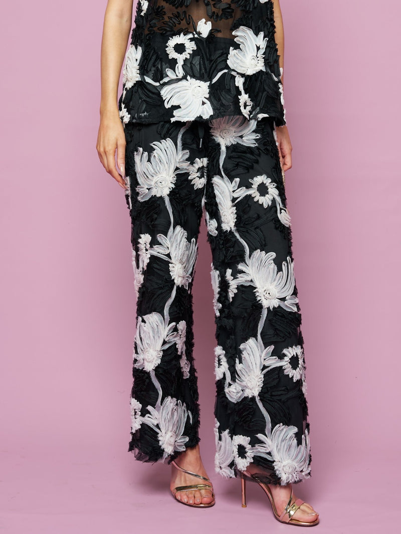 A person wears Social Queen's Contrast Floral Sheer Wide-Leg Pants and matching top with metallic heeled sandals, posing against a pink backdrop.