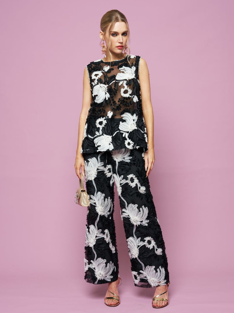 A woman poses against a pink background wearing Social Queen's Contrast Floral Sheer Wide-Leg Pants with bold white floral prints, paired with a sleeveless black top, a small purse, and open-toe heels.