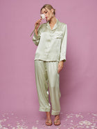 A woman poses against a pink background in the Social Queen Contrast-Trim Silk Pajama Set, holding a flower near her face with petals scattered around her feet.