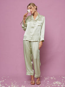 A woman poses against a pink background in the Social Queen Contrast-Trim Silk Pajama Set, holding a flower near her face with petals scattered around her feet.