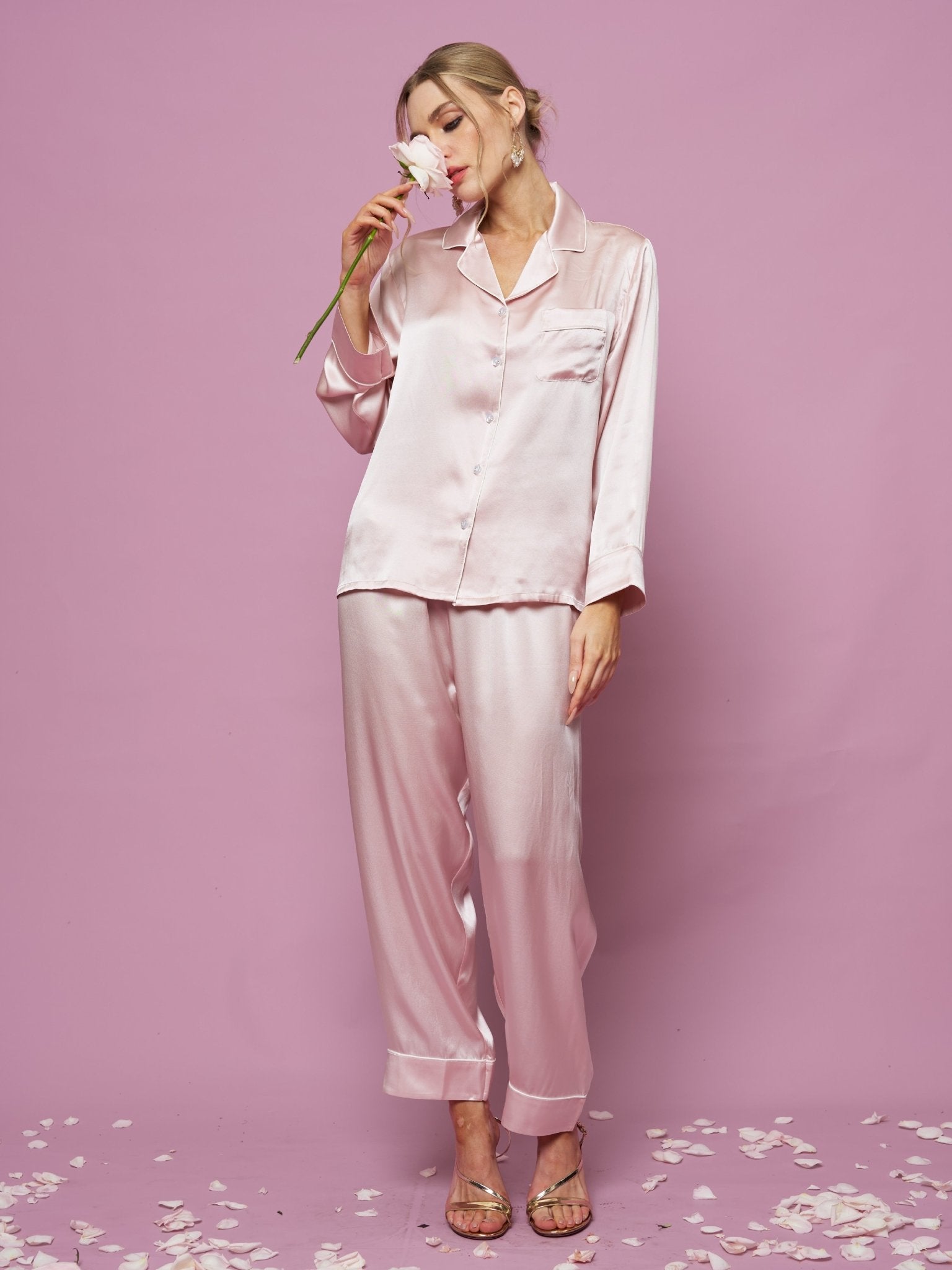 A woman wears the Social Queen Contrast-Trim Silk Pajama Set, standing against a pink backdrop and holding a flower to her face, with petals scattered around her feet.