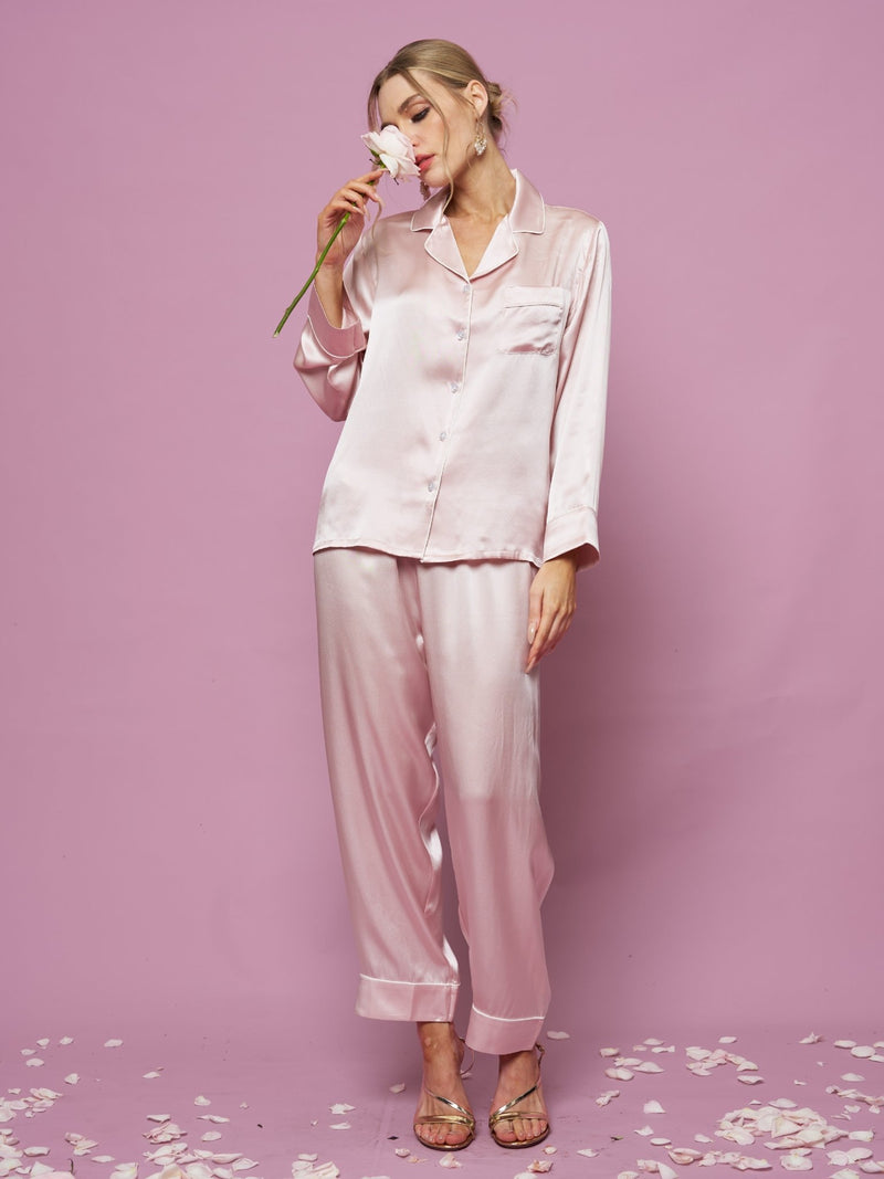 A woman wears the Social Queen Contrast-Trim Silk Pajama Set, standing against a pink backdrop and holding a flower to her face, with petals scattered around her feet.