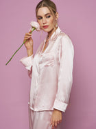 A woman wearing the Social Queen Contrast-Trim Silk Pajama Set holds a light pink rose near her face, standing against a solid pink background.
