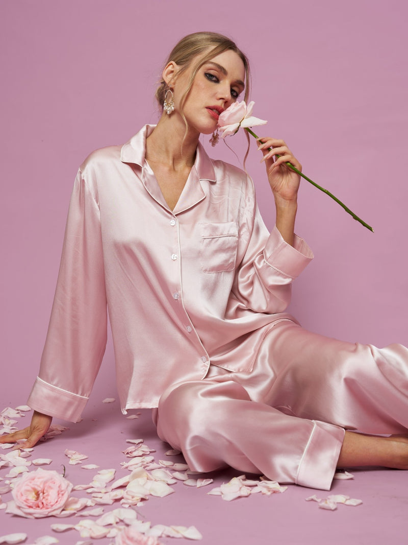 A woman wearing the Social Queen Contrast-Trim Silk Pajama Set sits on the floor against a pink background, holding a pink rose near her face with rose petals scattered around her.