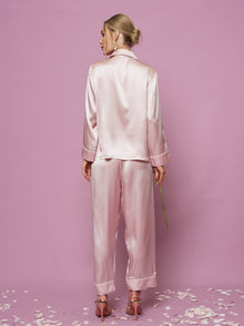A woman in a Social Queen Contrast-Trim Silk Pajama Set and high-heeled sandals stands facing away, holding a flower stem, with rose petals on the pink floor.