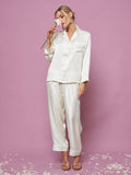 A person in the Social Queen Contrast-Trim Silk Pajama Set and sandals stands on a pink floor scattered with flower petals, holding a pink flower near their face against a pink background.
