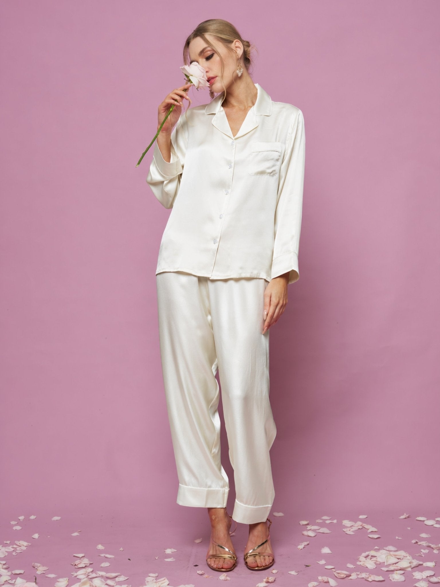 A person in the Social Queen Contrast-Trim Silk Pajama Set and sandals stands on a pink floor scattered with flower petals, holding a pink flower near their face against a pink background.
