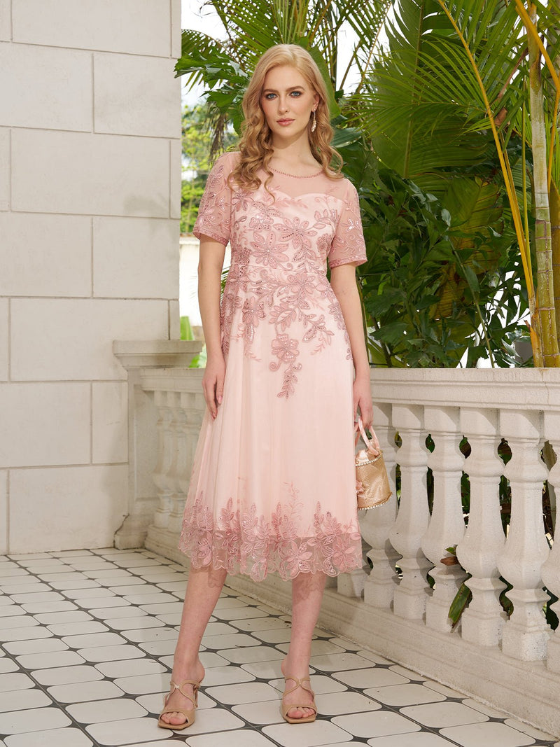 A woman stands on a tiled balcony with greenery, holding a purse and wearing the Social Queen Cordonnet Floral Sequin Shimmer Tulle Dress—a light pink, knee-length A-line dress with floral embroidery and short sleeves.