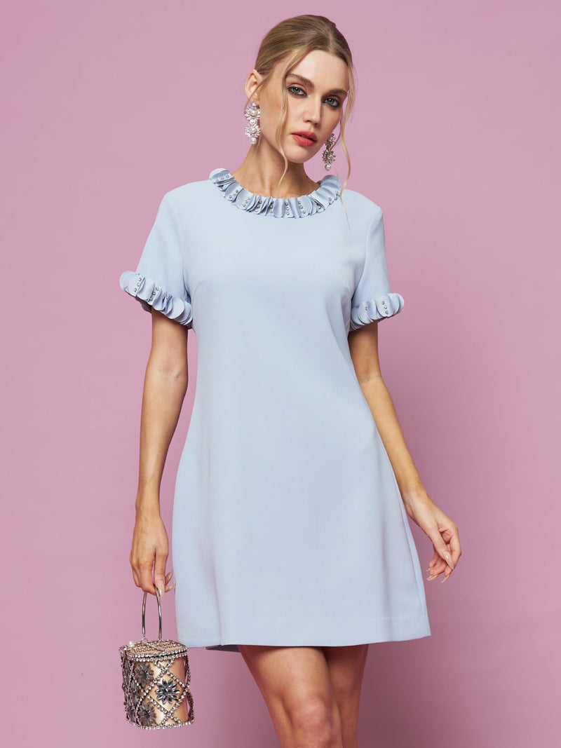 A woman in the Social Queen Crystal Beaded Trim Dress stands against a pink background, holding a small snake-print handbag.