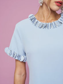 A woman wears the Social Queen Crystal Beaded Trim Dress, featuring a light blue A-line silhouette with ruffled, crystal-beaded trim on the sleeves and neckline, set against a pink background.