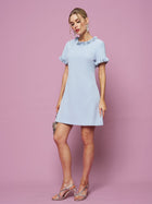 A woman in the Social Queen Crystal Beaded Trim Dress, featuring light blue satin with ruffled sleeves and collar, silver heels, and earrings, poses against a pink background.