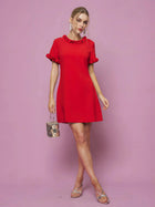 Woman in the Social Queen Crystal Beaded Trim Dress, a short red satin piece with ruffled sleeves and hem in an A-line silhouette, holds a small handbag while standing against a pink background.