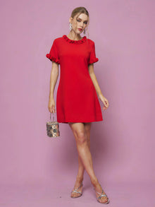 Woman in the Social Queen Crystal Beaded Trim Dress, a short red satin piece with ruffled sleeves and hem in an A-line silhouette, holds a small handbag while standing against a pink background.