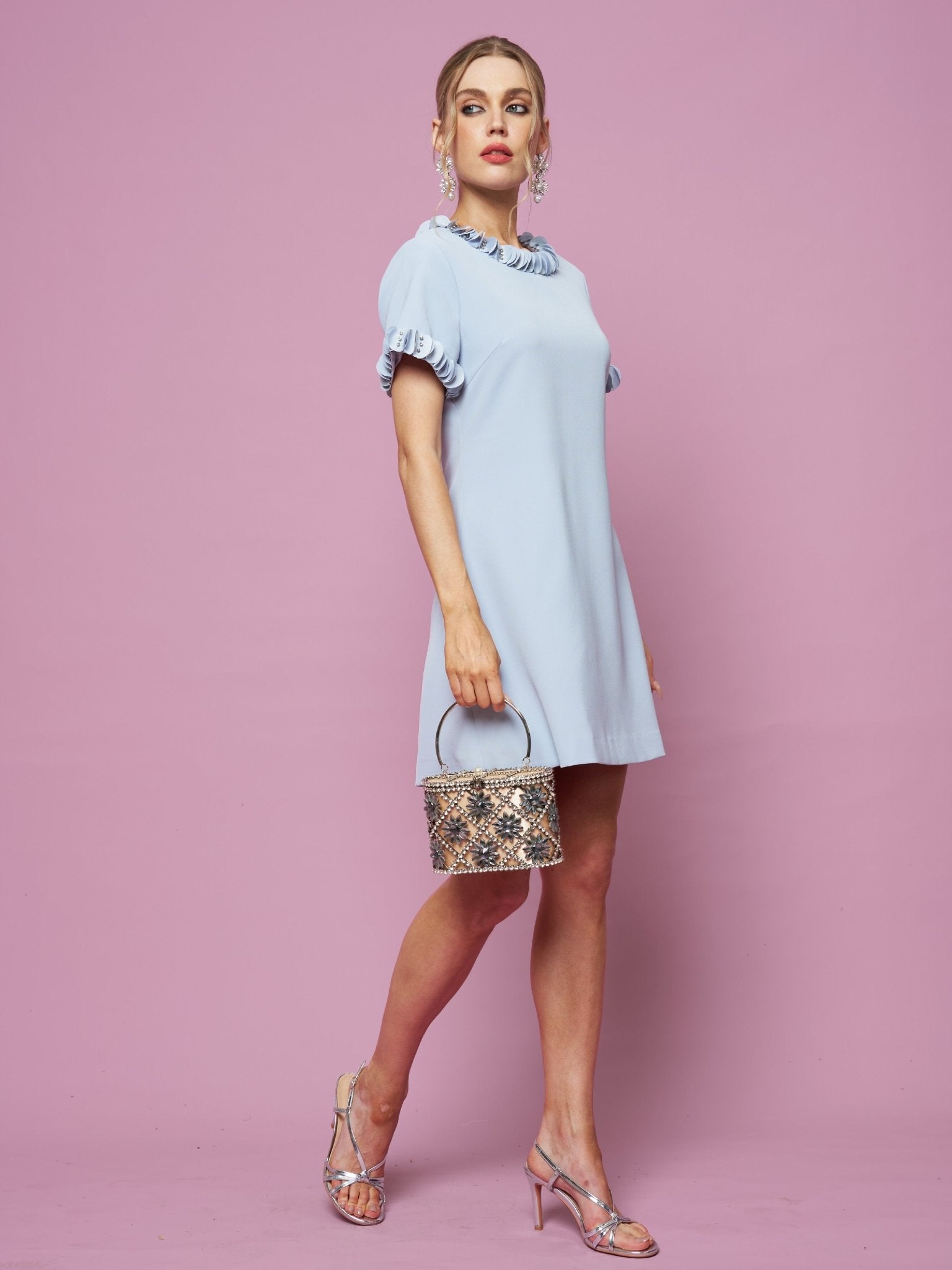 A woman in the Social Queen Crystal Beaded Trim Dress stands against a pink background, holding a small snakeskin handbag and wearing silver heels.