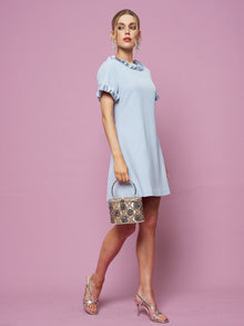 A woman in the Social Queen Crystal Beaded Trim Dress stands against a pink background, holding a small snakeskin handbag and wearing silver heels.