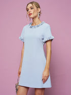 A woman in the Social Queen Crystal Beaded Trim Dress, a light blue satin A-line with short ruffled sleeves and collar, holds a small purse against a pink background.