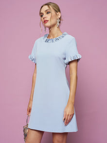 A woman in the Social Queen Crystal Beaded Trim Dress, a light blue satin A-line with short ruffled sleeves and collar, holds a small purse against a pink background.