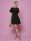 A woman wears the Social Queen Crystal Beaded Trim Dress, a short black satin style with ruffled sleeves and collar, against a pink background. She pairs it with a metallic handbag and silver sandals. The A-line silhouette is flattering.