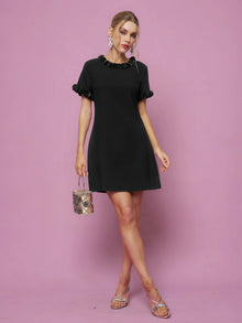 A woman wears the Social Queen Crystal Beaded Trim Dress, a short black satin style with ruffled sleeves and collar, against a pink background. She pairs it with a metallic handbag and silver sandals. The A-line silhouette is flattering.