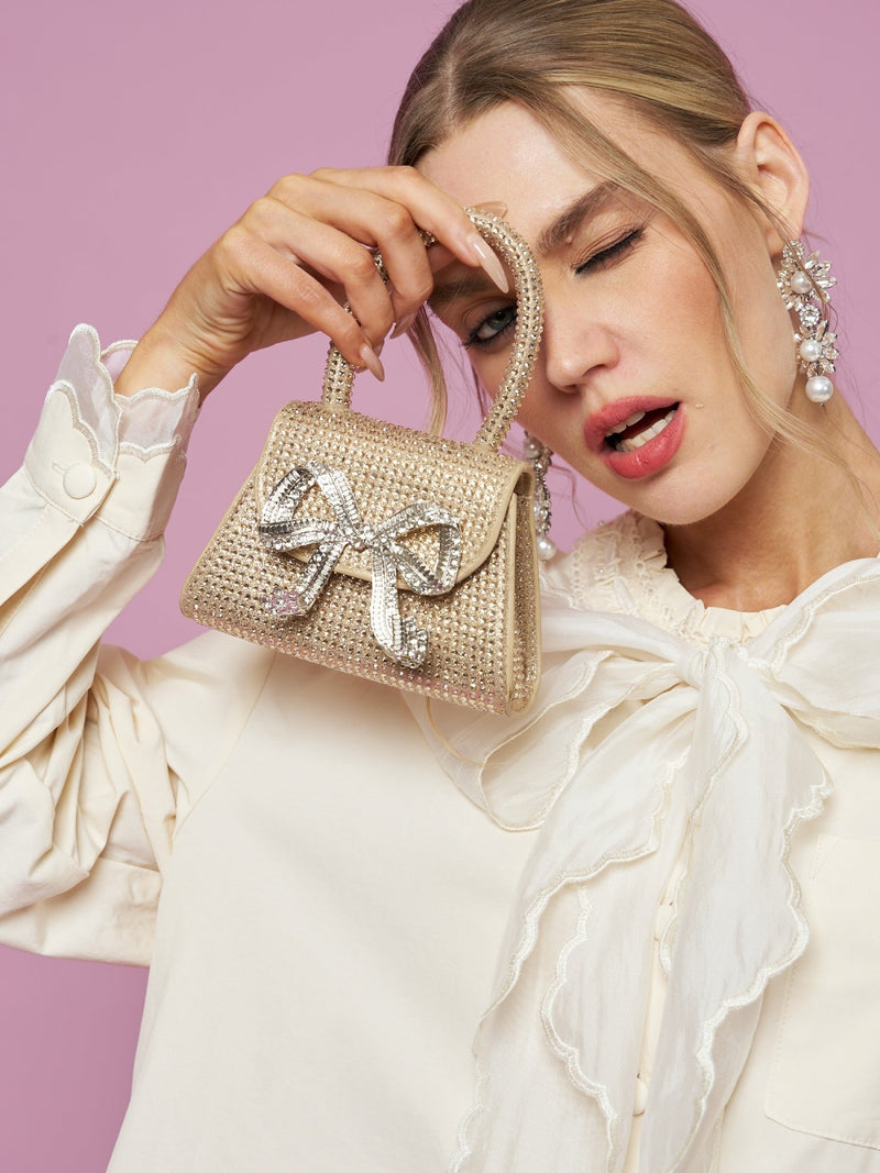 A woman in a cream blouse poses against a pink background, winking and holding the Social Queen Crystal-Embellished Bow Mini Bag.