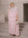 Wearing Social Queen’s Crystal-Embellished Cape Dress, a person stands on stairs with a beaded clutch, statement earrings, and heels.