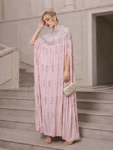 Wearing Social Queen’s Crystal-Embellished Cape Dress, a person stands on stairs with a beaded clutch, statement earrings, and heels.