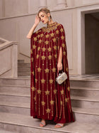 A woman stands on stairs wearing the Social Queen Crystal-Embellished Cape Dress, paired with silver sandals, statement earrings, and holding a silver clutch.