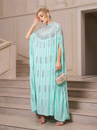 A woman stands on stairs wearing the Social Queen Crystal-Embellished Cape Dress with sequin detailing, paired with silver earrings, heels, and a small ornate clutch.