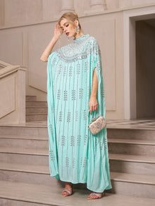 A woman stands on stairs wearing the Social Queen Crystal-Embellished Cape Dress with sequin detailing, paired with silver earrings, heels, and a small ornate clutch.
