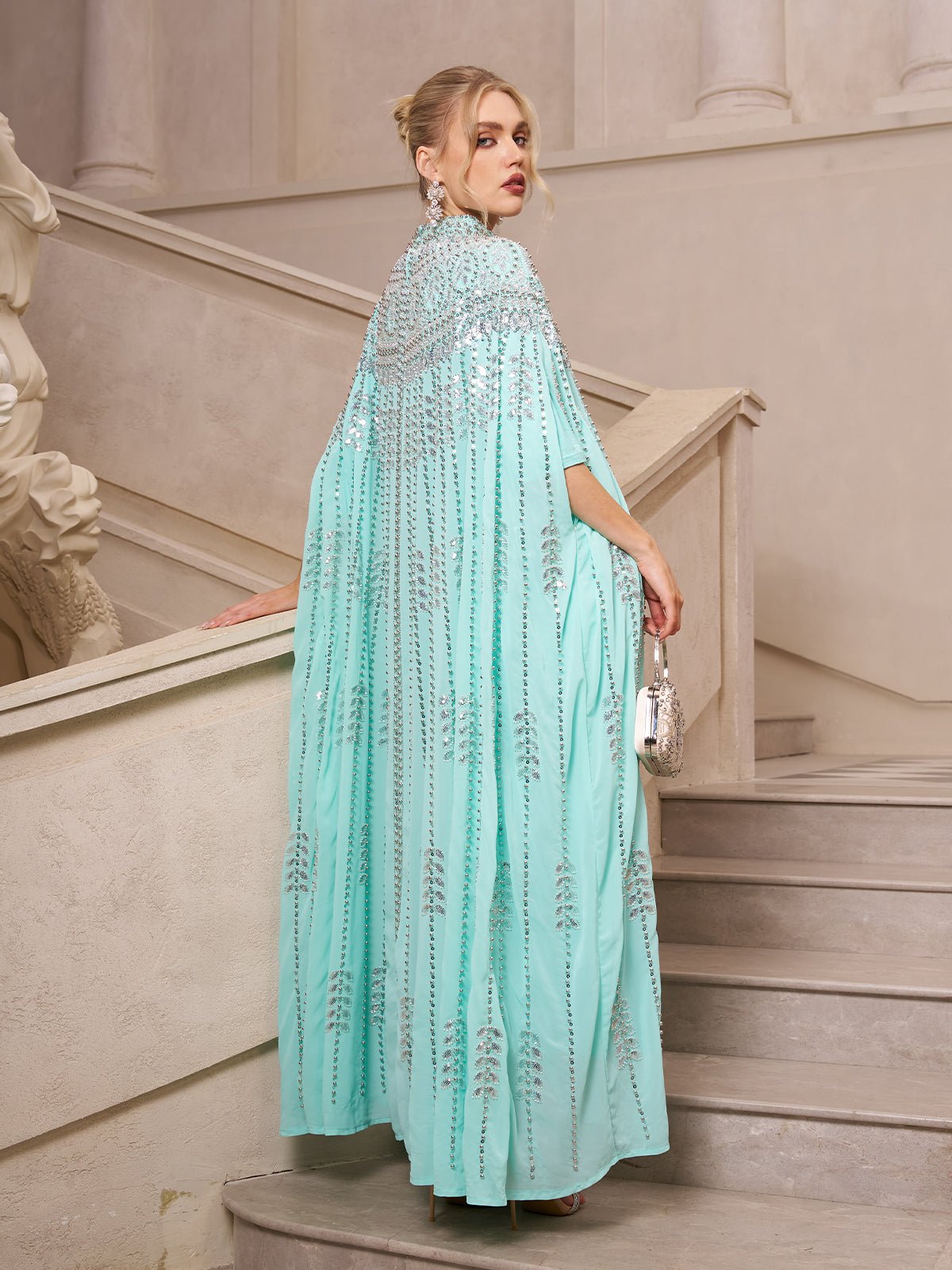 A woman in the Social Queen Crystal-Embellished Cape Dress stands on a staircase, looking over her shoulder and holding a small silver purse.
